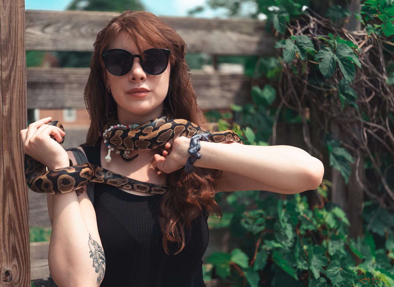 A woman standing outdoors wearing sunglasses and holding a ball python.